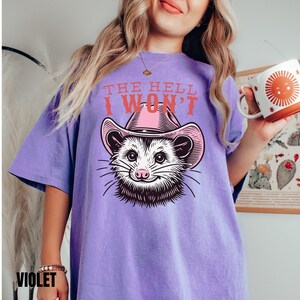 Pu&ograve; includere: Maglietta viola con una grafica di un opossum che indossa un cappello da cowboy rosa. Il testo "THE HELL I WON'T" &egrave; stampato sulla maglietta.
