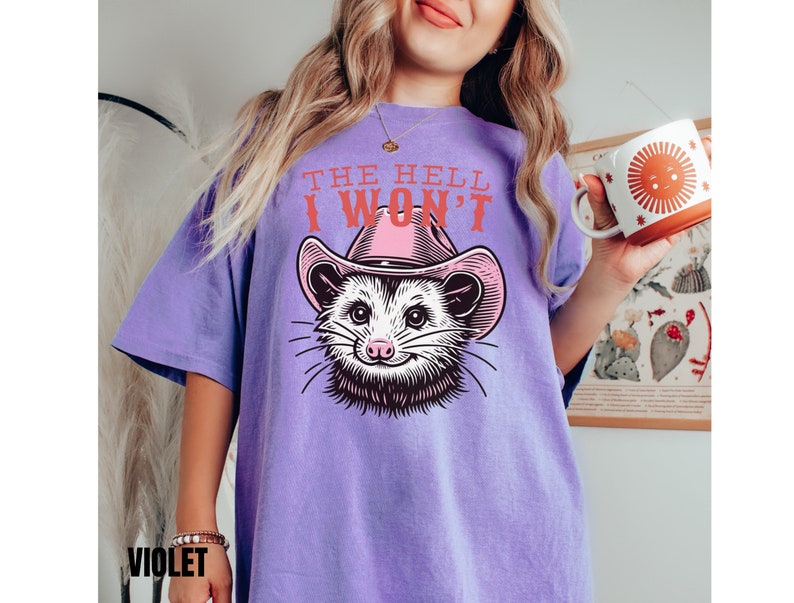 Pu&ograve; includere: Maglietta viola con una grafica di un opossum che indossa un cappello da cowboy rosa. Il testo "THE HELL I WON'T" &egrave; stampato sulla maglietta.