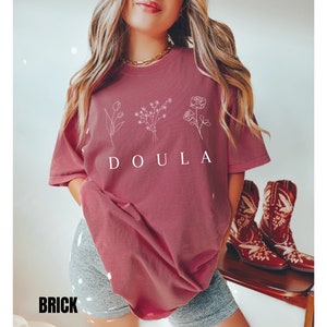 May include: A maroon t-shirt with a white graphic of flowers and the word "DOULA" printed on the front.