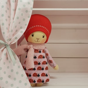 May include: A handmade doll with a red hat, pink dress with ladybug print, and a pink scarf. The doll is standing in front of a white shelf with a pink and green polka dot curtain.