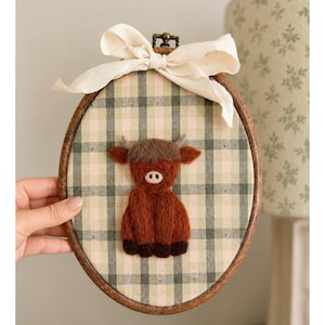 May include: An oval wooden embroidery hoop containing a fabric background with a green and beige plaid pattern. A needle-felted brown Highland cow figure is centred on the fabric. A cream-coloured fabric ribbon is tied in a bow at the top.