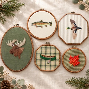 May include: Five embroidery hoops featuring nature-themed designs: an oval hoop with a brown moose head, a round hoop with a fish, an octagonal hoop with a flying duck, an octagonal hoop with a green canoe and paddle, and a round hoop with a red maple leaf.