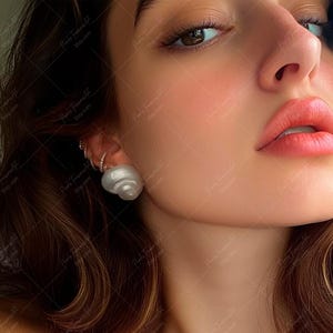 Puede incluir: Primer plano de la cara de una persona con un pendiente de concha blanca. La persona tiene el pelo largo y castaño y lleva maquillaje.