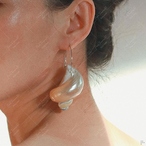 May include: A pair of distinctive earrings with a large, pearlescent, seashell-shaped design. The earrings hang from thin, silver-coloured hoops. The seashell elements show a gradient of colours, from cream to light orange.