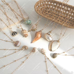 Puede incluir: Una colección de collares dorados, cada uno con un colgante de concha marina único. Las conchas varían en tamaño, forma y color, incluyendo conchas blancas, marrones y negras. Los collares están dispuestos sobre una superficie blanca, con una cesta tejida en el fondo.