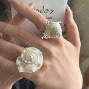 Könnte beinhalten: Zwei Muschelschmuckringe. Ein Ring zeigt eine große, flache, weiße Muschel mit Spiralmuster, der andere eine kleinere, konische Muschel. Beide Ringe haben silberfarbene Metallakzente und sind an einer Hand getragen.