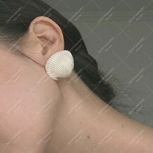 May include: A white seashell earring, a large scallop shell, worn on an ear. The earring's textured surface is visible. The background shows a person's shoulder and hair. The Etsy shop name is visible in the background.