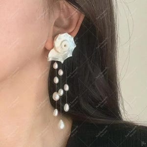 Puede incluir: Pendientes blancos en forma de concha con perlas blancas colgantes. Los pendientes tienen un diseño detallado y en capas que se asemeja a una concha, con múltiples hilos de perlas que caen en cascada hacia abajo. Las perlas varían en tamaño.