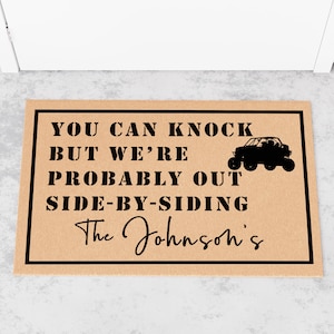 May include: A brown doormat with black text that reads "You can knock but we're probably out side-by-siding The Johnson's". A black silhouette of a side-by-side vehicle is to the right of the text.