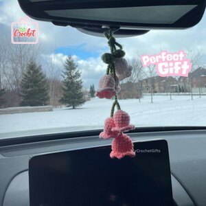 May include: A pink and green crocheted flower garland hanging from a car's rearview mirror. The garland features multiple pink bell-shaped flowers and green leaves. The text "Perfect Gift" is visible in the background.