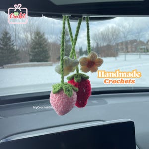 May include: Handmade crocheted strawberry car charms with green leaves and pink and red flowers. The charms are hanging from the rearview mirror of a car. The text "Handmade Crochets" is visible in the image.