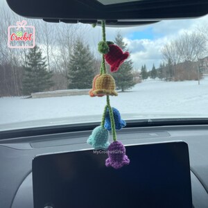 May include: A rainbow-coloured crocheted flower garland hanging from a car's rearview mirror. The garland features five flowers in different colours: red, yellow, blue, green, and purple. The flowers are crocheted with a delicate stitch and have a bell-like shape. The garland is a cheerful and whimsical addition to any car.