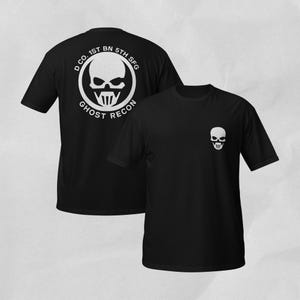 May include: A black short-sleeved t-shirt featuring a white skull graphic on the left chest. The back displays a large circular emblem with a skull and the text D CO. 1ST BN 5TH SFG GHOST RECON printed in white around the perimeter of the circle.