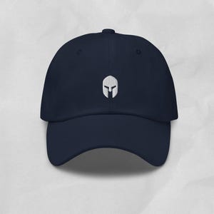 Könnte beinhalten: Eine marineblaue Baseballkappe mit einem weißen, gestickten Spartanerhelm-Symbol in der Mitte der Vorderseite. Die Kappe verfügt über eine strukturierte Krone, einen gebogenen Schirm und einen Knopf an der Oberseite für ein schlichtes Design.