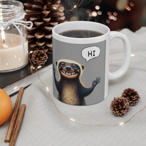 May include: White ceramic mug with a gray background featuring a cartoon honey badger with a toothy grin and the word "HI" in a speech bubble. The mug is filled with coffee.