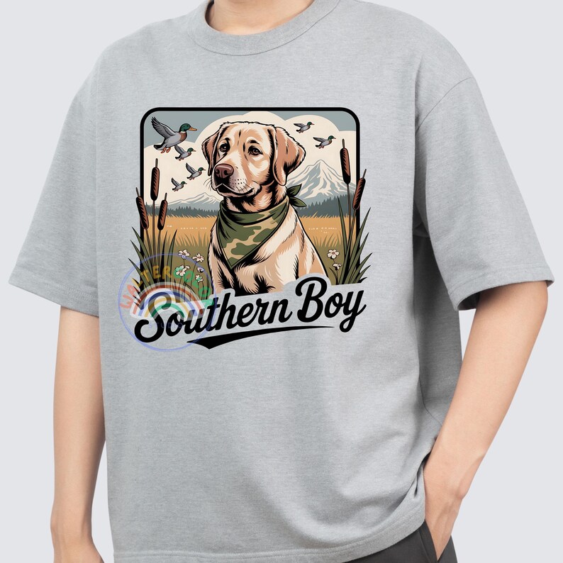 Op de afbeelding: Lichtgrijs t-shirt met een afbeelding van een gele Labrador Retriever met een groene bandana. De hond staat tegen een achtergrond van een veld, bergen en vliegende eenden. De tekst "Southern Boy" staat eronder.