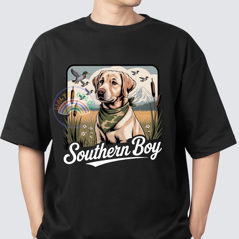 Op de afbeelding: Zwart t-shirt met een Labrador Retriever met een groene bandana en de tekst "Southern Boy". Het ontwerp bevat eenden, een berg en hoog gras, waardoor een ontwerp met een natuurthema ontstaat.