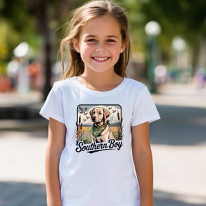 Op de afbeelding: Wit T-shirt met een afbeelding van een gele Labrador Retriever met een bandana, tegen een landschap met vogels en riet. De tekst "Southern Boy" staat eronder. Het T-shirt wordt door een jong meisje gedragen.