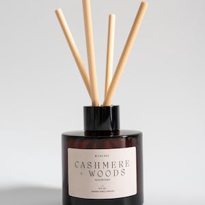 May include: A brown glass reed diffuser with a black cap and wooden reeds. The label reads "Cashmere + Woods Reed Diffuser" with the text "100 ml Handmade in West Yorkshire". This home fragrance product is designed to scent a room.