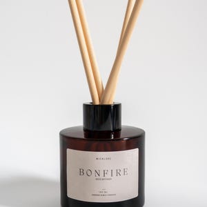 May include: A dark brown glass reed diffuser with a black cap and wooden reeds. The label reads "BONFIRE". The diffuser is designed to release fragrance into the air. The bottle contains 100 ml of liquid.