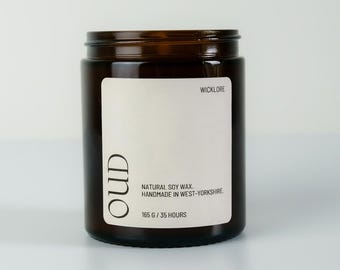 May include: A brown glass jar candle with a white label. The label reads "OUD" vertically, with "WICKLORE" at the top. The label also states "NATURAL SOY WAX. HANDMADE IN WEST-YORKSHIRE. 165 G / 35 HOURS."