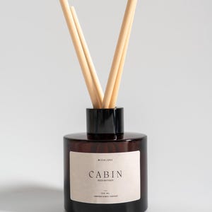 May include: A brown glass reed diffuser with a black cap and light-coloured reeds. The label reads "CABIN" and "REED DIFFUSER". The bottle contains 100 ml of liquid.