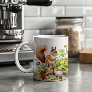 May include: White ceramic mug with a colorful illustration of a red squirrel sitting in a flower pot. The squirrel is surrounded by green plants and red flowers. The mug has a handle on the left side.