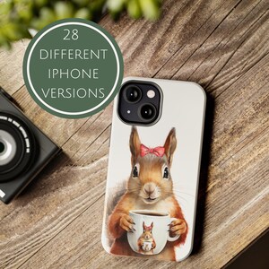 Könnte beinhalten: Weißes iPhone-Case mit einer Aquarellillustration eines Eichhörnchens mit roter Schleife, das eine Kaffeetasse hält. Die Tasse hat eine kleinere Eichhörnchen-Illustration. Das Case befindet sich neben einer Kamera und einem grünen Kreis mit dem Text "28 VERSCHIEDENE IPHONE VERSIONEN".