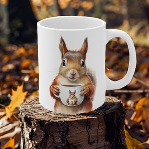 May include: White ceramic mug with a brown squirrel holding a smaller white mug with a brown squirrel inside. The squirrel is holding the mug with both paws and has a happy expression on its face.