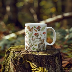 May include: White ceramic mug with a woodland design featuring squirrels, mushrooms, and ferns. The mug has a handle and is sitting on a tree stump.