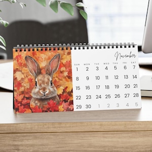 May include: A desk calendar with a watercolour illustration of a brown rabbit amidst autumn leaves. The calendar shows the month of November with dates from 1 to 30. The calendar is spiral-bound and rests on a wooden surface.