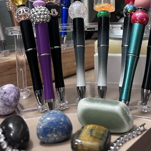 May include: A collection of eight decorative pens with various colored tops and glitter accents. The pens are displayed on a clear plastic stand. The stand is surrounded by several small, colorful stones.