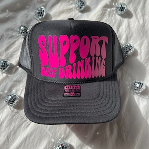 Könnte beinhalten: Schwarze Trucker-Kappe mit einem pinkfarbenen "Support Day Drinking"-Aufdruck in einer Retro-Schrift.