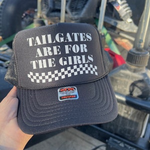 May include: Black trucker hat with the text "TAILGATES ARE FOR THE GIRLS" in white block letters. The hat has a black mesh back and a white and black checkered pattern. A sticker on the front reads "WORLD FAMOUS FOAM FRONT".