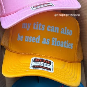 May include: A stack of trucker hats in pink, yellow, and blue. The pink hat has the text "Blowjobs are real jobs" in orange. The yellow hat says "my tits can also be used as floaties" in blue. The blue hat says "MY GUMMY JUST KICKED IN" in pink.