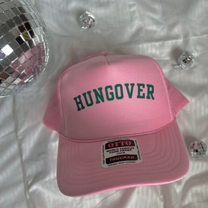 May include: A pink trucker hat with a green "HUNGOVER" logo on the front. The hat has a black mesh back and a white label with the text "OTTO WORLD FAMOUS FOAM FRONT OSFM TRUCKER DURABLE SNAP CLOSURE".