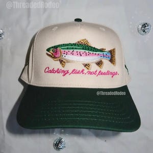 May include: A beige and green baseball cap featuring an embroidered rainbow trout. The cap has a green brim and the text "Catching fish, not feelings" in pink script. The brand name "ThreadedRodeo" is visible.