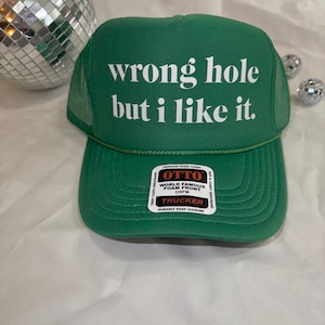 May include: Green trucker hat with the text "wrong hole but I like it" in white. The hat has a mesh back and a curved brim. The hat also has a tag that says "OTTO WORLD FAMOUS FOAM FRONT TRUCKER".