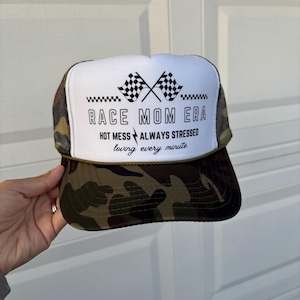 May include: A trucker hat with a white front panel and a camouflage print on the bill and back. The hat features the text "RACE MOM ERA" with crossed checkered flags and the phrases "HOT MESS ALWAYS STRESSED loving every minute."