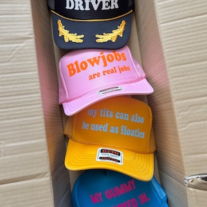 May include: A selection of trucker hats in a cardboard box. The hats are black, pink, yellow, and blue, each with a different phrase in bold text. The black hat says "TOPLESS DRIVER" and has gold accents. The pink hat says "BLOWJOBS are real jobs".