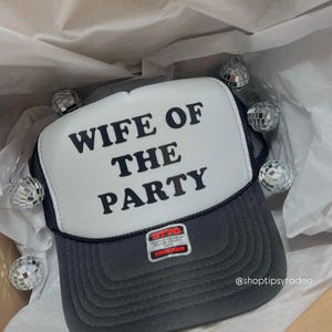 Pode incluir: Um chapéu de caminhoneiro branco com texto preto que diz "Wife of the Party". O chapéu tem uma parte de trás de malha preta e uma aba preta. O chapéu está sobre um fundo de papel de seda branco.