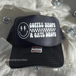 May include: Black trucker hat with white text that reads "Coffee Shops & Gate Drops". The hat also features a white smiley face with lightning bolts for eyes.