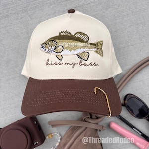 Peut inclure: Casquette de baseball avec visière marron et couronne crème. Le chapeau présente un motif de poisson brodé et l'inscription "kiss my bass". Un hameçon doré est fixé à la visière.