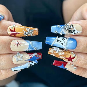 Op de afbeelding: Een set lange, vierkante kunstnagels. Elke nagel heeft een uniek ontwerp, waaronder ster- en bloemaccenten, koeienprint en een mix van rode, blauwe en bruine kleuren. Sommige nagels hebben zilveren versieringen.