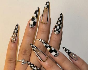 May include: Long, stiletto-shaped artificial nails with a black and white checkered pattern, clear sections, and gold and rhinestone accents. Several rings adorn the fingers.