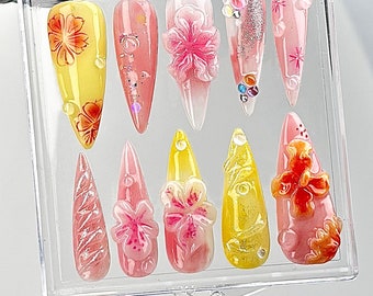 Puede incluir: Una vitrina de plástico transparente con 10 uñas postizas. Las uñas están decoradas con diferentes diseños florales en tonos rosa, amarillo y naranja.