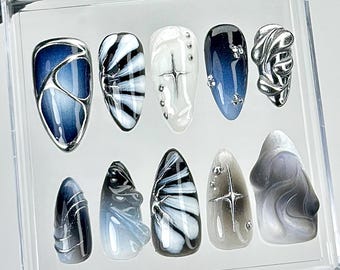 以下が含まれることがあります： 透明なプラスチックケースに入った10枚のネイルチップセット。アーモンド型とスティレット型が含まれています。深いブルーのグラデーション、白黒のマーブル模様、シルバーのメタリックなアクセント、立体的な3Dテクスチャー、星のモチーフが施されています。