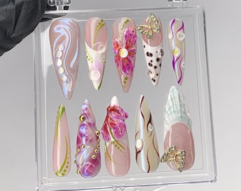 May include: A set of ten long, almond-shaped artificial nails in a clear plastic case. The nails feature various designs, including floral patterns, swirls, and butterfly accents. Colors include pink, white, gold, and purple.