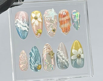 May include: A set of ten artificial nails in a clear plastic case. The nails feature various designs, including ocean waves, seashells, and floral accents. Colors include blue, pink, yellow, and iridescent finishes. Some nails have 3D embellishments.