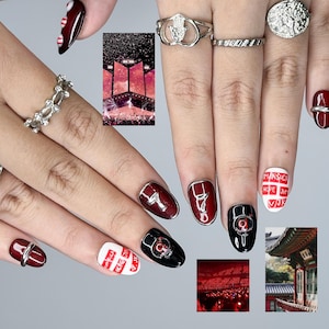 May include: Close-up of manicured nails with diverse designs. Some nails are burgundy with silver accents, others are black with silver embellishments, and some are white with red text. Silver rings adorn the fingers.